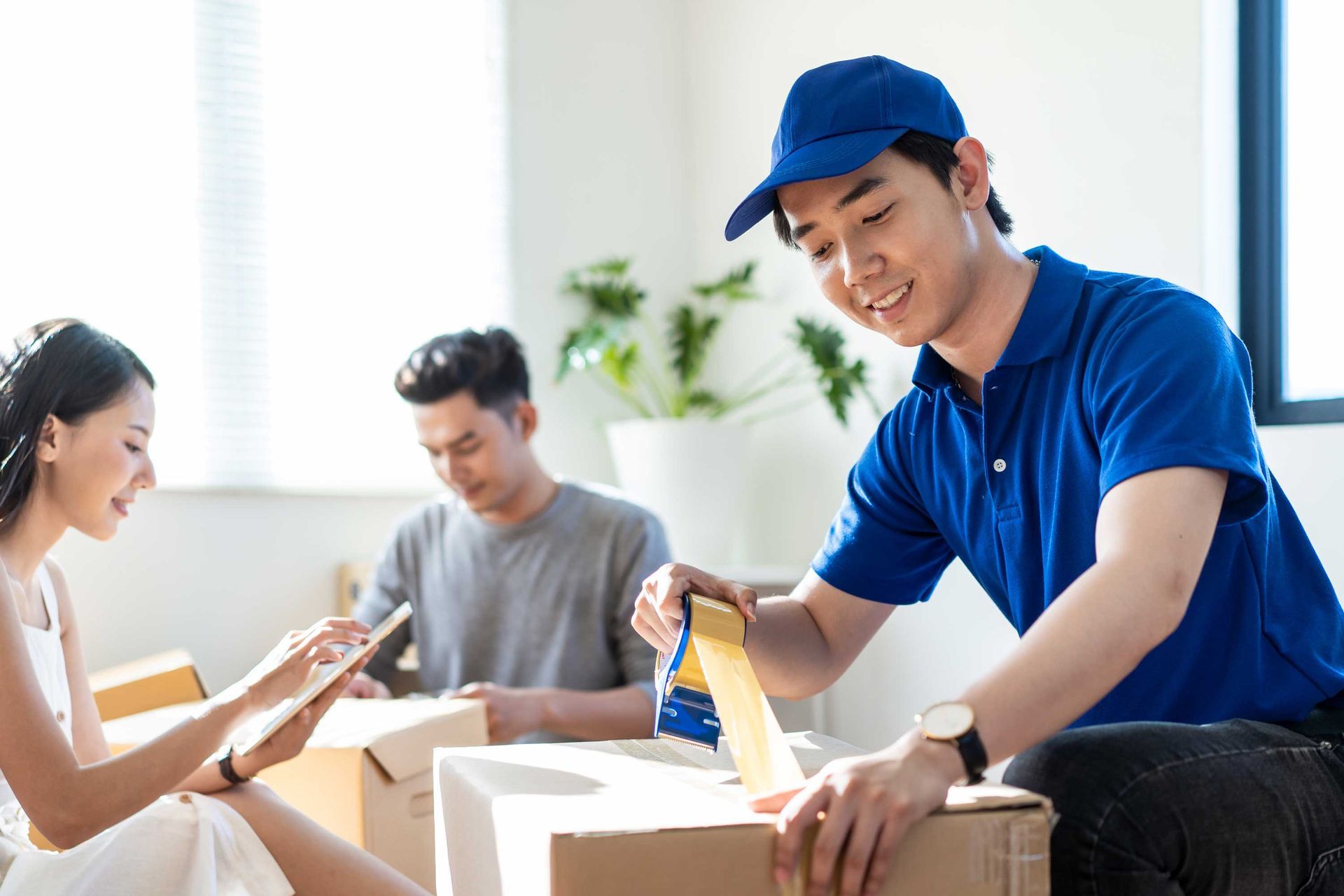Delivery person taping a box while a couple packs items indoors.