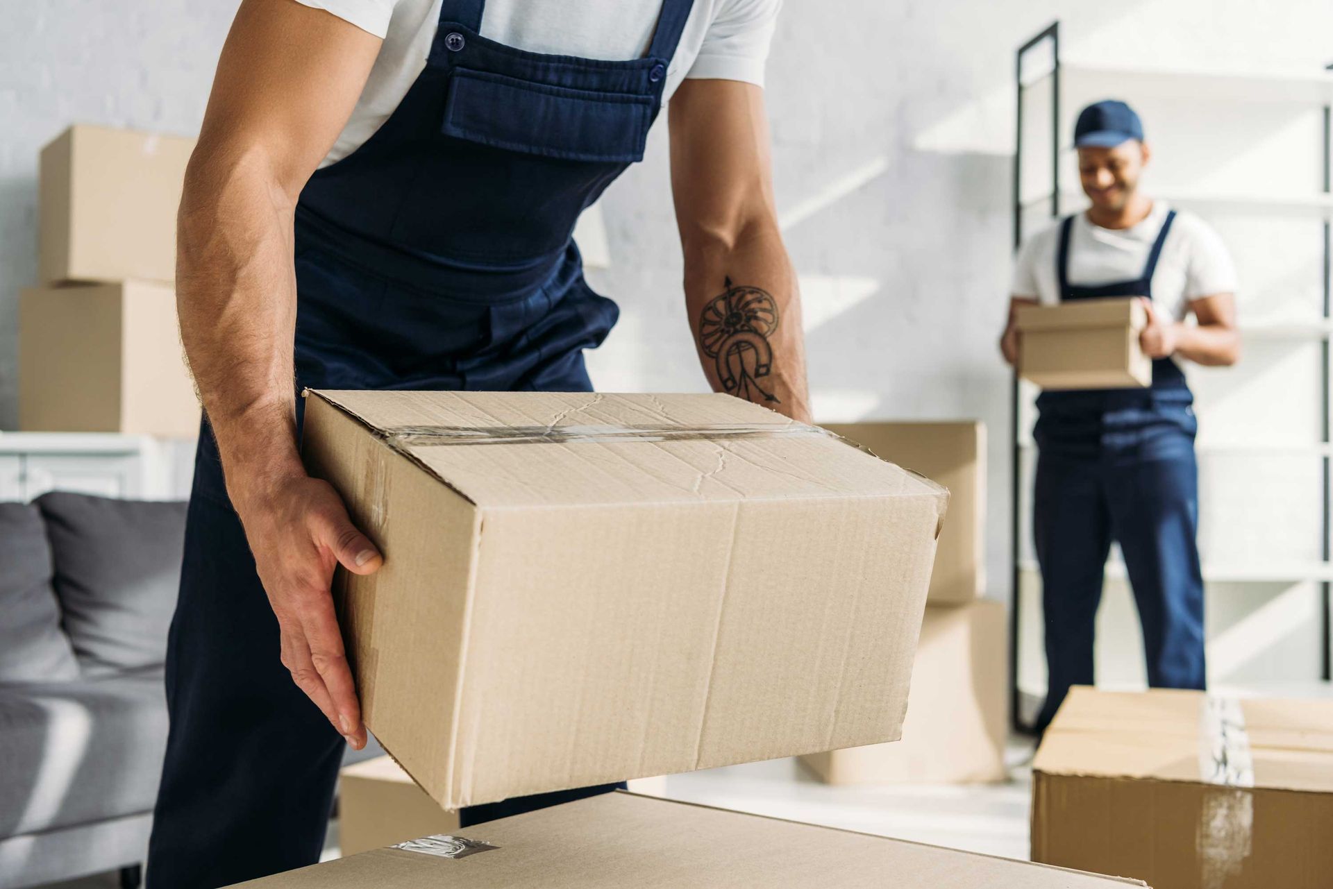 Two movers in blue coveralls lifting cardboard boxes in a room, ready to pack for relocation.