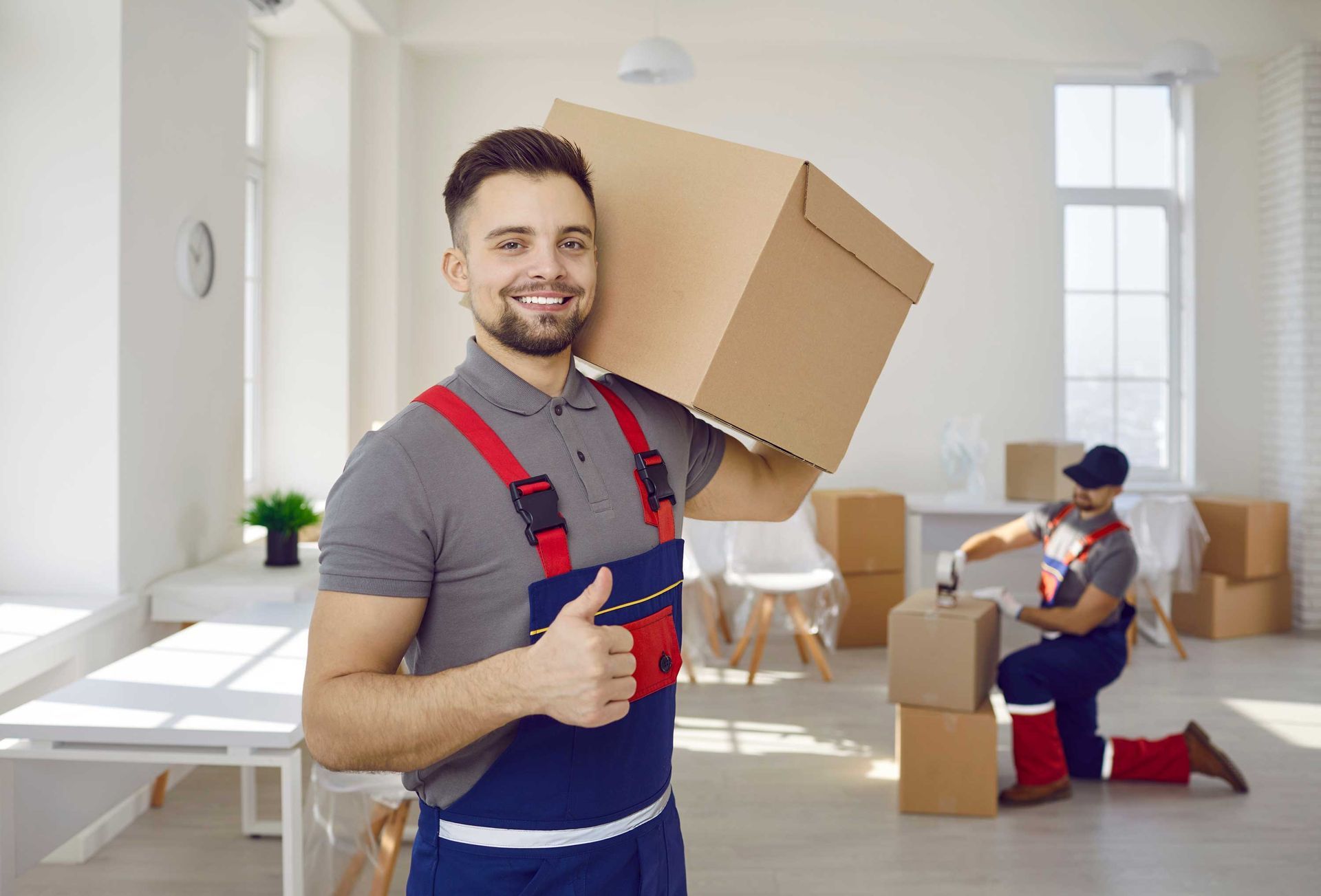 Man in overalls carrying a box on his shoulder, giving a thumbs-up. Another person packs boxes. Bright room.
