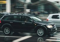 Black SUV driving on a city street, blurred background of buildings and other cars.