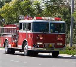 Red and white fire engine driving on a road with emergency lights flashing, trees in background.