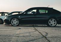 Black car with custom wheels on a paved surface, with tire tracks and two other cars in the background.