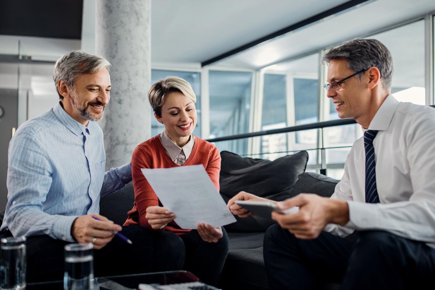Three colleagues review documents together on a sofa in a bright office lounge.