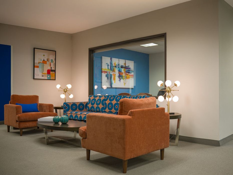 Modern lounge with orange armchairs, patterned sofa, glass tables, and abstract wall art.