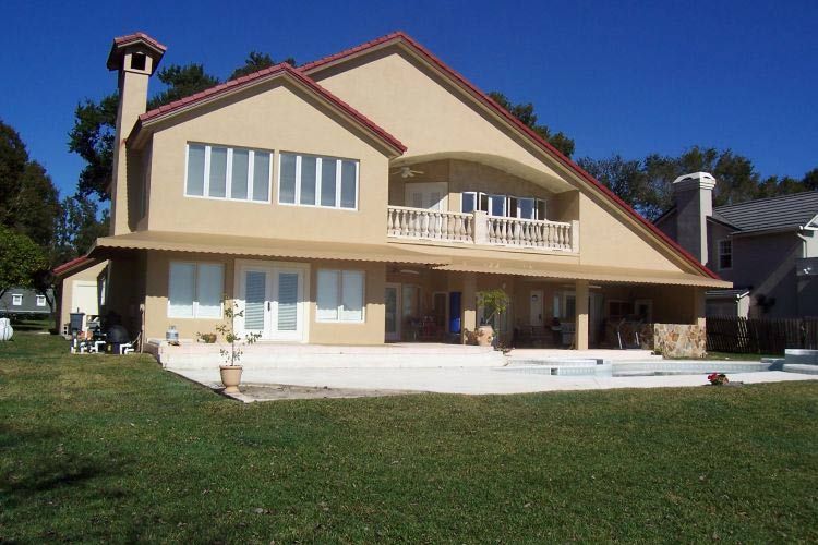 Two-story stucco house with red roof trim and a balcony, overlooking a grassy yard on a sunny day.