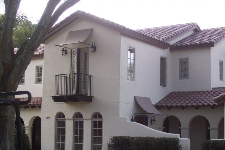 White stucco multi-story building with red tile roof, arched windows, small balcony, and brown awning.