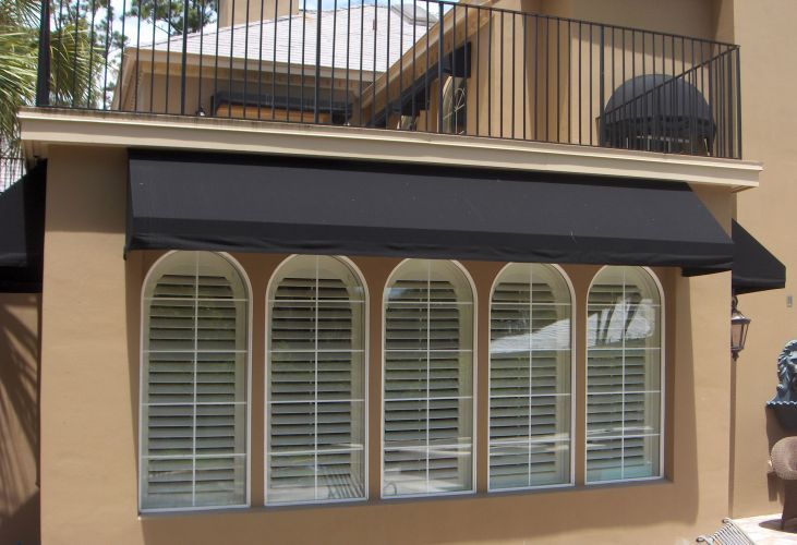 Black awning above arched windows, beige building. Balcony above, sunny outdoor setting.