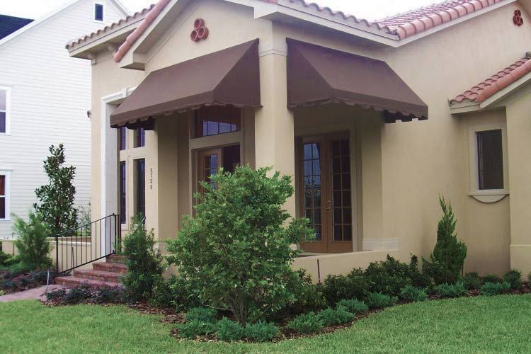 Tan stucco house with brown awnings over windows, green landscaping, and red tile roof.