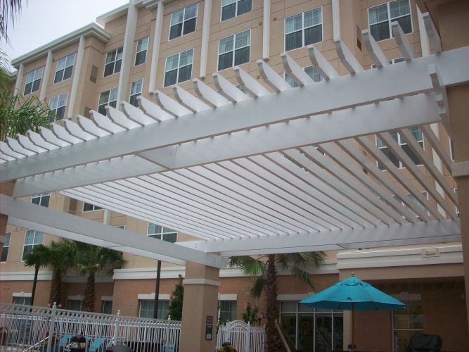 White pergola over a pool area; building and blue umbrella in the background.