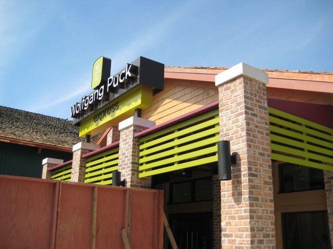 Wolfgang Puck Express restaurant entrance with brick columns, yellow signage, and green accents.