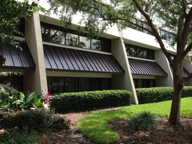 Two-story modern building with dark awning and large windows, surrounded by greenery and a tree.