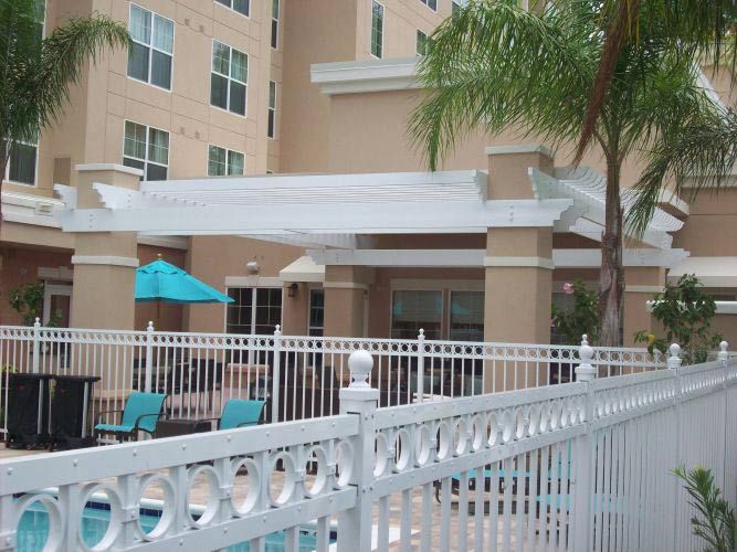 Hotel entrance with white pergola, pool, and white fence. Blue chairs and umbrella near the pool.