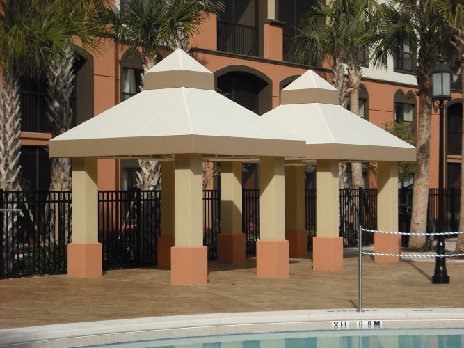 Two beige and tan pavilions near a pool, with a building and palm trees in the background.