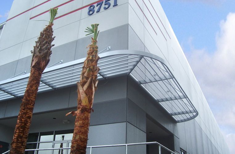 Building exterior with gray and white panels, curved metal awning, palm trees, and the number 8751.