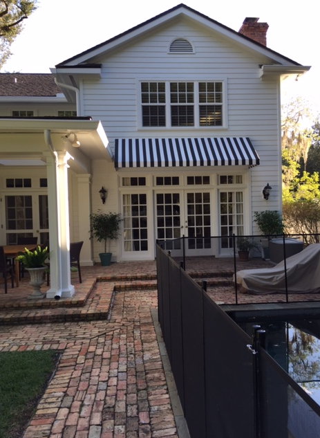 White house with brick pathway, French doors under a blue-striped awning, and pool area.