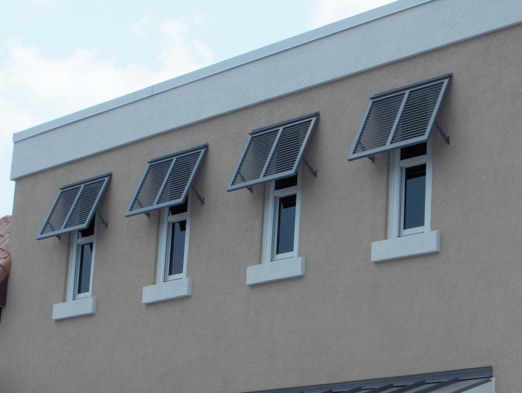 Four windows with open, slatted awnings on a light tan building under a cloudy sky.