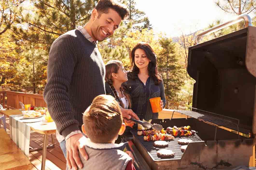 Family grilling outdoors: parents and children smiling, burgers and skewers on grill, fall setting.