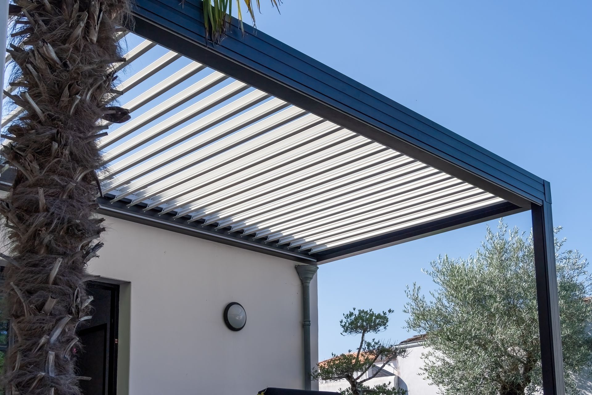 Modern slatted pergola attached to a house, with palm trees and blue sky overhead. Modern slatted pergola attached to a house, with palm trees and blue sky overhead.