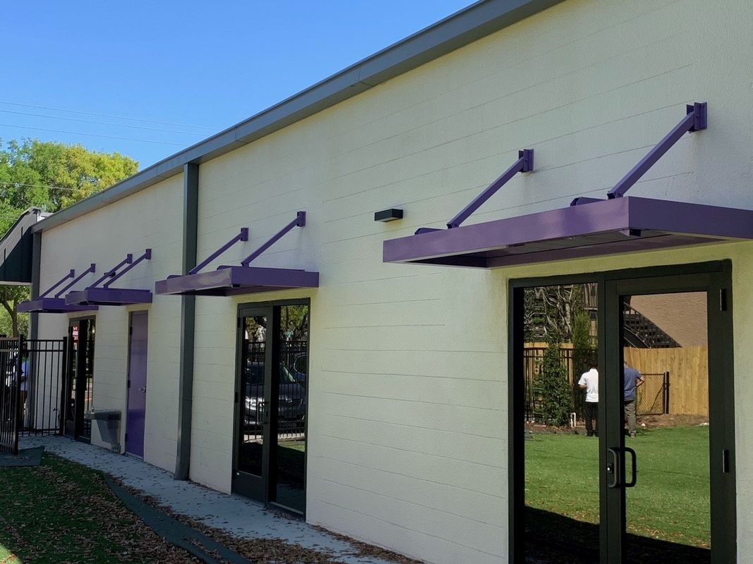 Exterior of a building with purple awnings over glass doors, against a white brick wall. Sunny day.