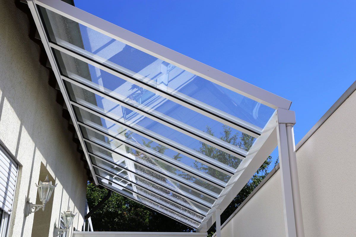 White-framed patio cover with clear panels against a blue sky, attached to a cream-colored wall.