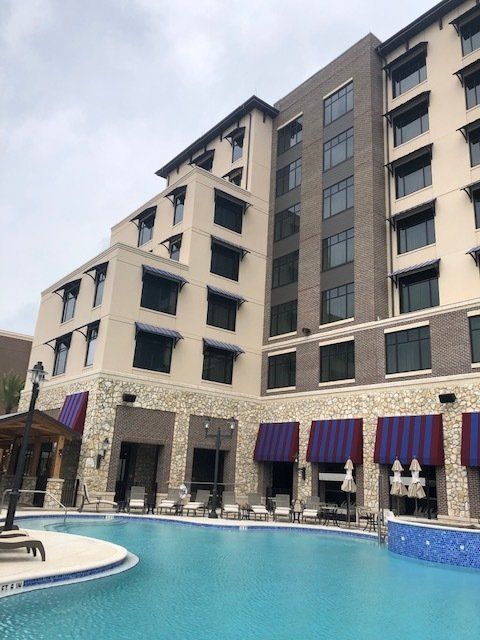A modern building with pool, awnings, and stone facade. Blue pool water reflects cloudy sky.
