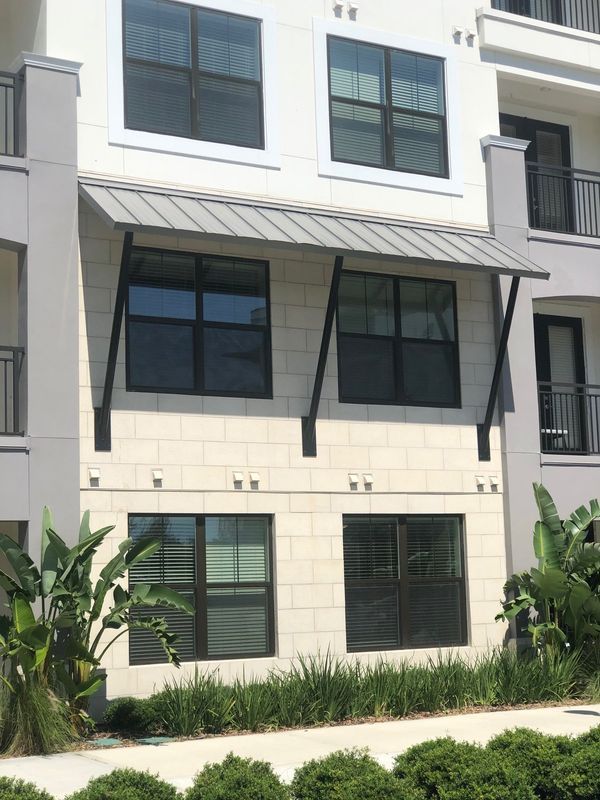 Building exterior with light stone facade, dark-framed windows, metal awning, and greenery.