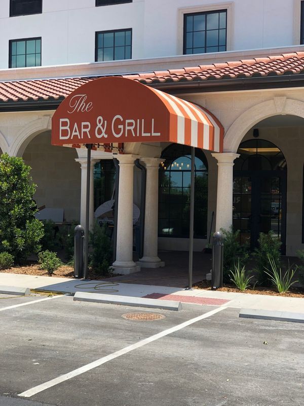 Entrance to The Bar & Grill under an orange and white striped awning, with white columns and arches, in front of a building.