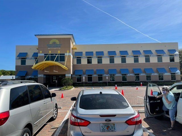 Two-story building with blue awnings, yellow accents, and a parking lot with cars. Woman exiting a car.