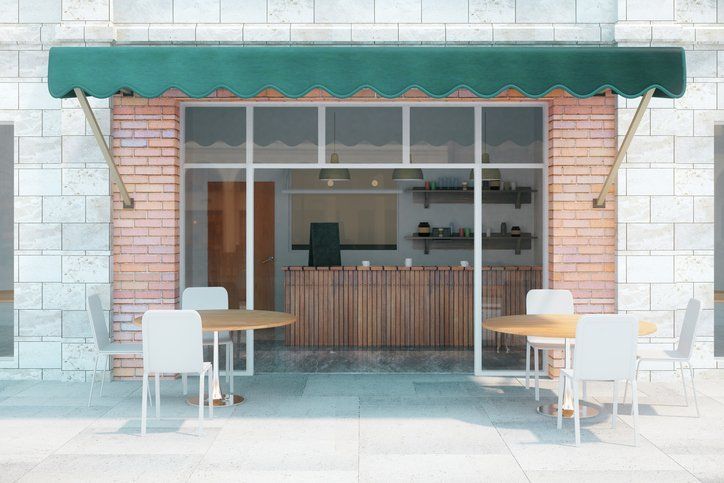 Cafe exterior with green awning, tables, and chairs. The front has large glass windows and a brick facade.