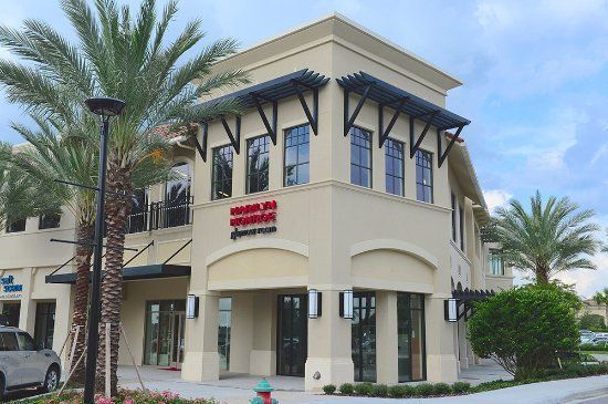Exterior of a beige, two-story building with black window frames, palm trees, and a red sign.
