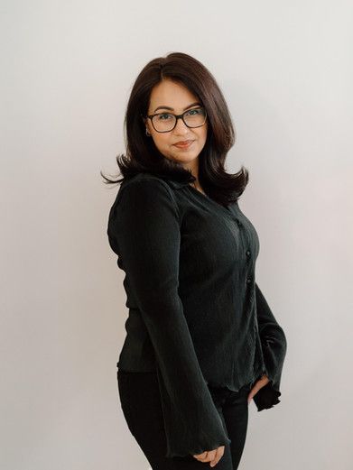 A woman wearing glasses and a black shirt is standing in front of a white wall.