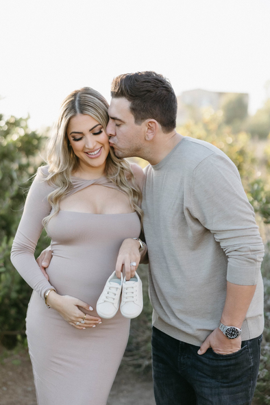 A man is kissing a pregnant woman on the cheek while she holds a pair of baby shoes.