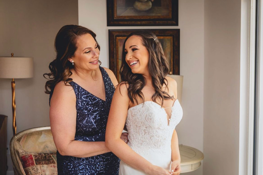 A bride and her mother are standing next to each other in front of a window.