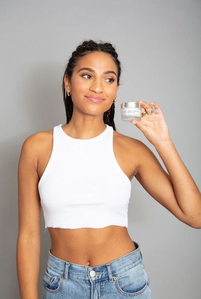A woman in a white crop top and blue jeans is holding a jar of cream.