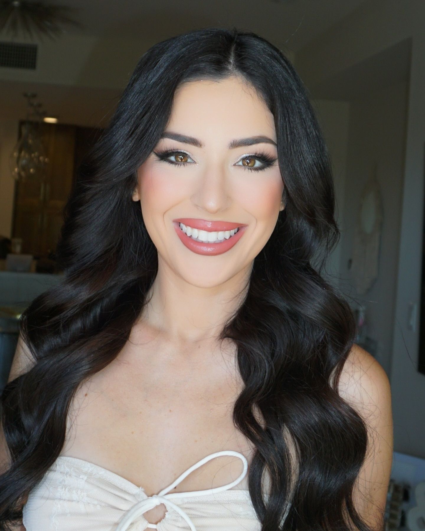 A woman with long dark hair is smiling and wearing a white top
