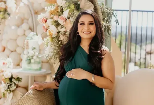 A pregnant woman in a green dress is holding her belly and smiling.