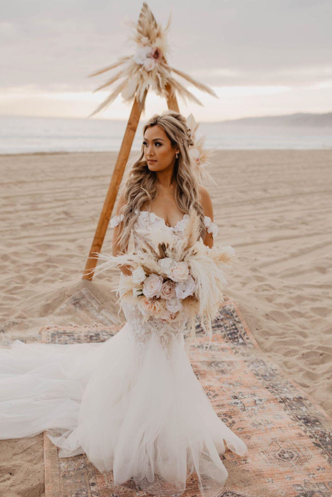 A bride in a wedding dress is standing on a beach holding a bouquet of flowers.