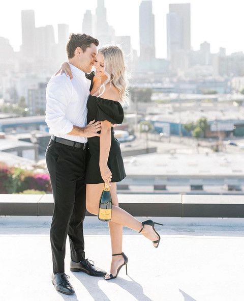 A man and a woman are standing next to each other on a rooftop holding a bottle of champagne.