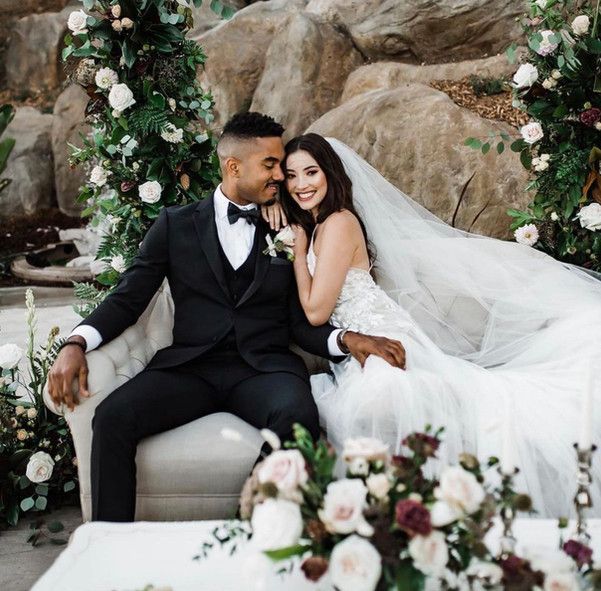 A bride and groom are sitting on a couch surrounded by flowers.
