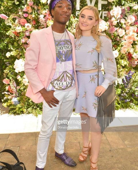 A man and a woman are standing next to each other in front of a wall of flowers.