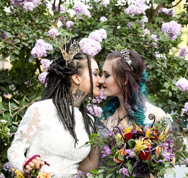 Two women are kissing in front of a bush with purple flowers.