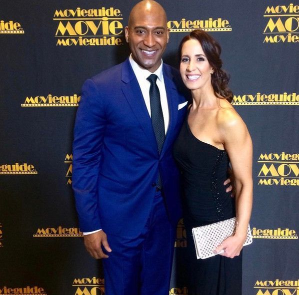 A man and woman pose in front of a wall that says movieguide