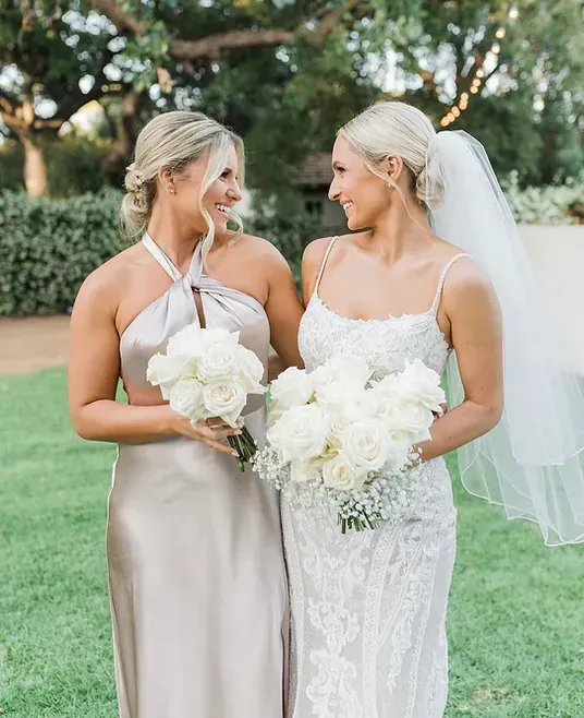 A bride and her bridesmaid are standing next to each other on a lush green field.