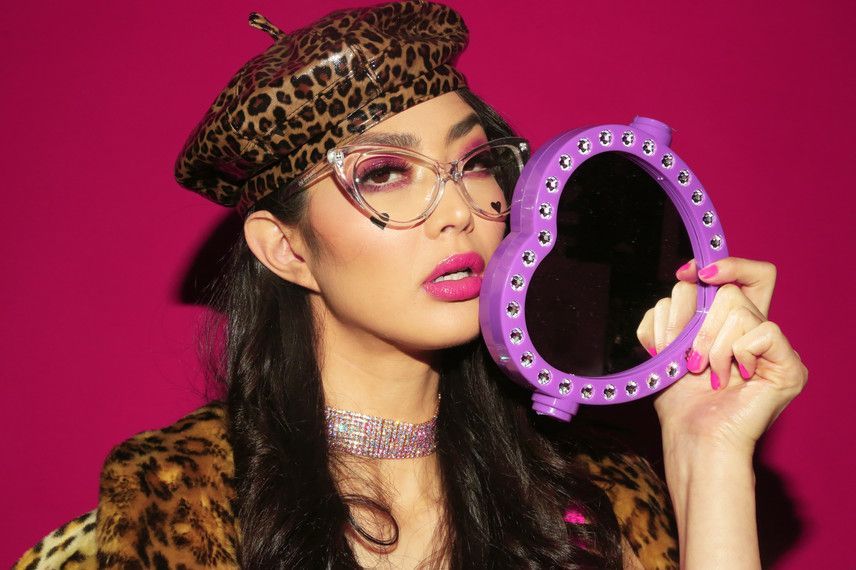 A woman wearing a leopard print hat and glasses is holding a purple heart shaped mirror.