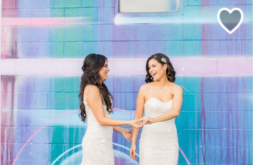 Two women in wedding dresses are holding hands in front of a colorful wall.