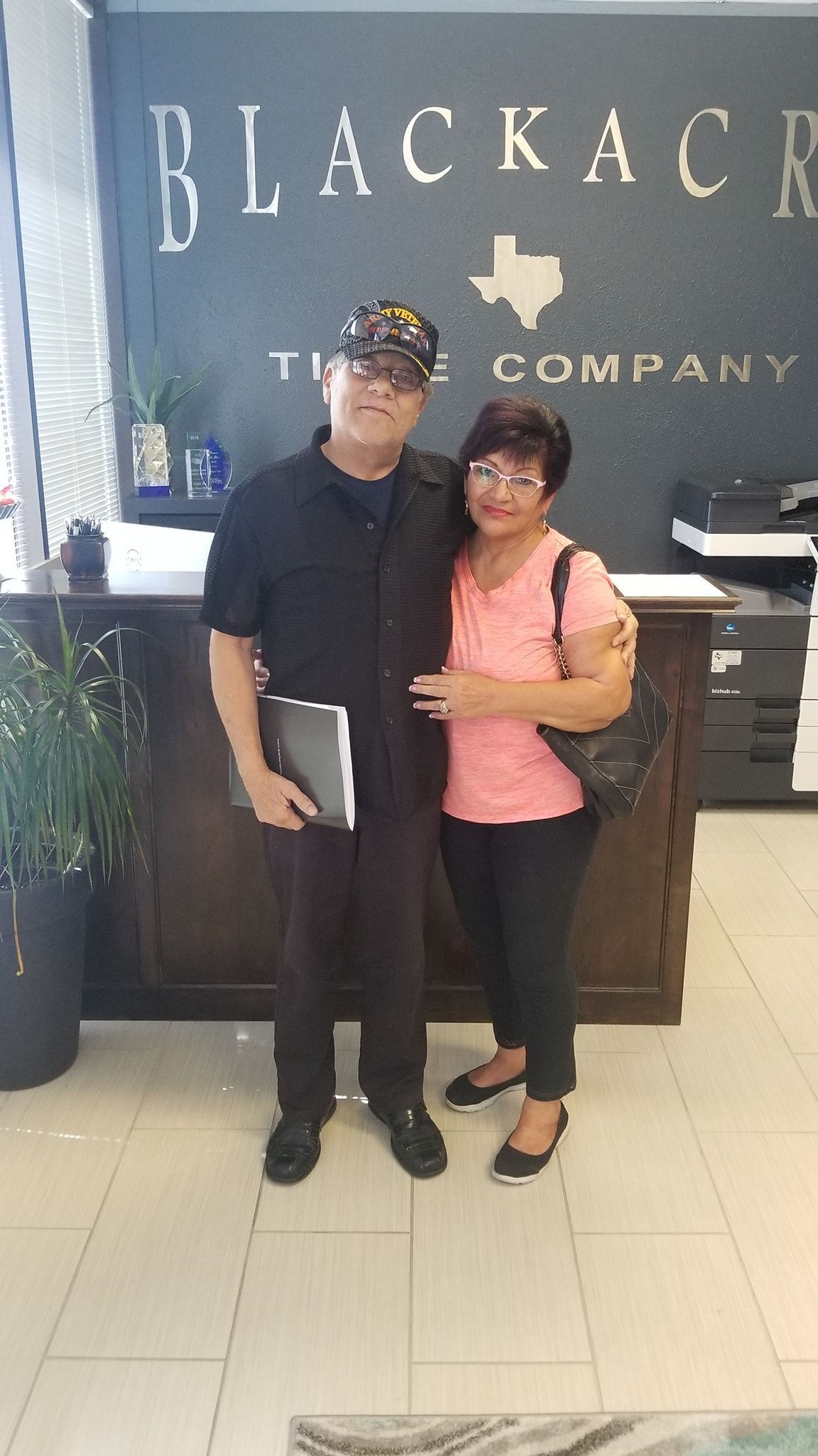 Couple poses in front of a Blackacre Tile Company sign. Man holds notebook. Woman wears pink shirt.
