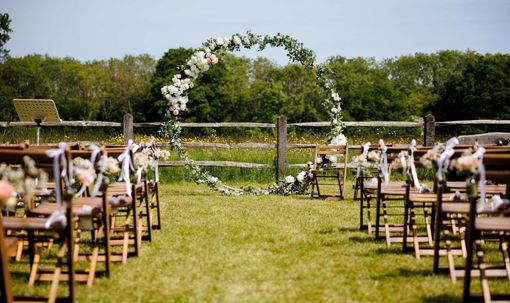 A row of wooden chairs are lined up in a grassy field for a wedding ceremony.
