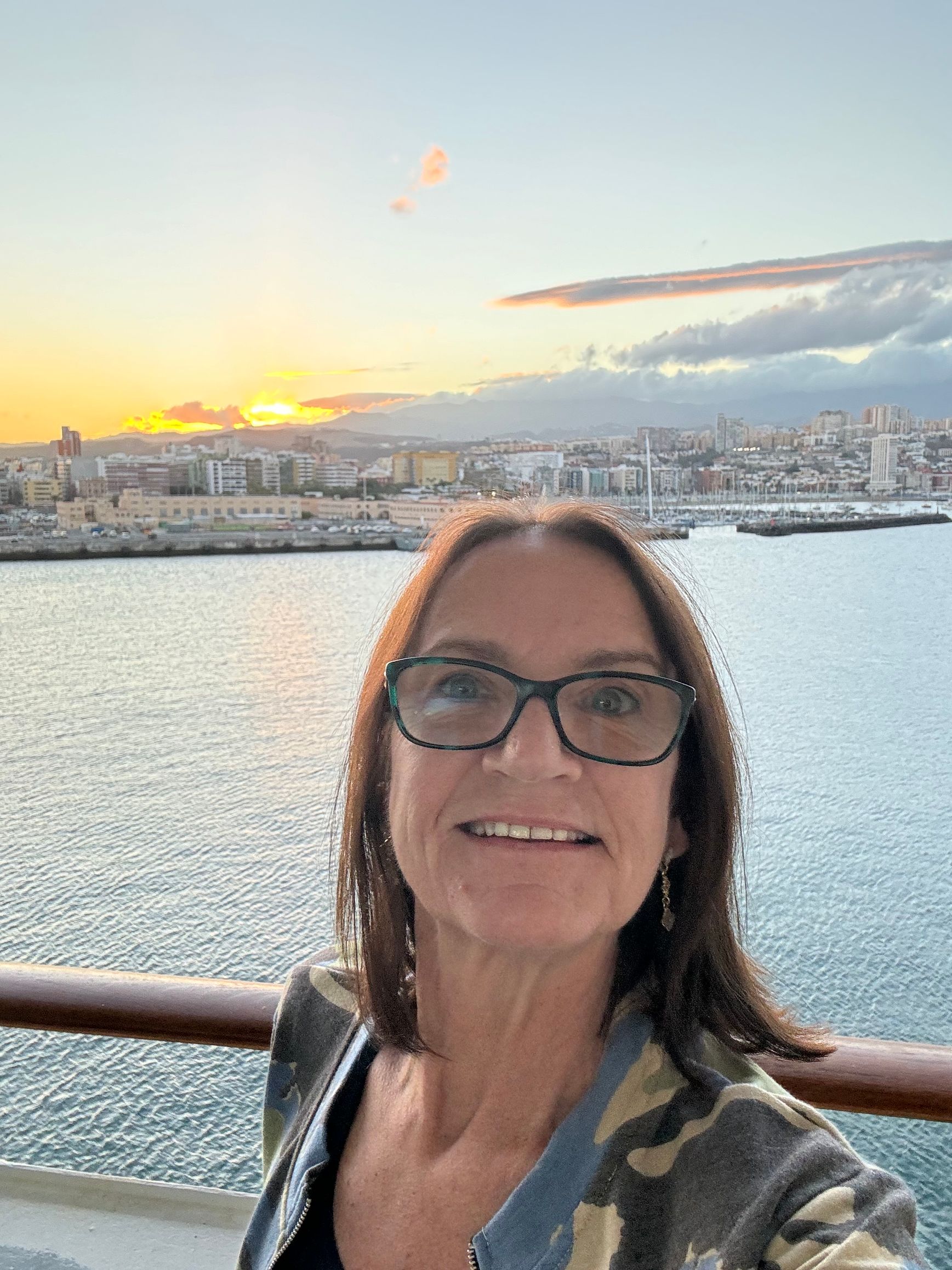 Danielle Webster  in glasses smiles on a cruise ship deck at sunset, overlooking a city skyline in Europe.
