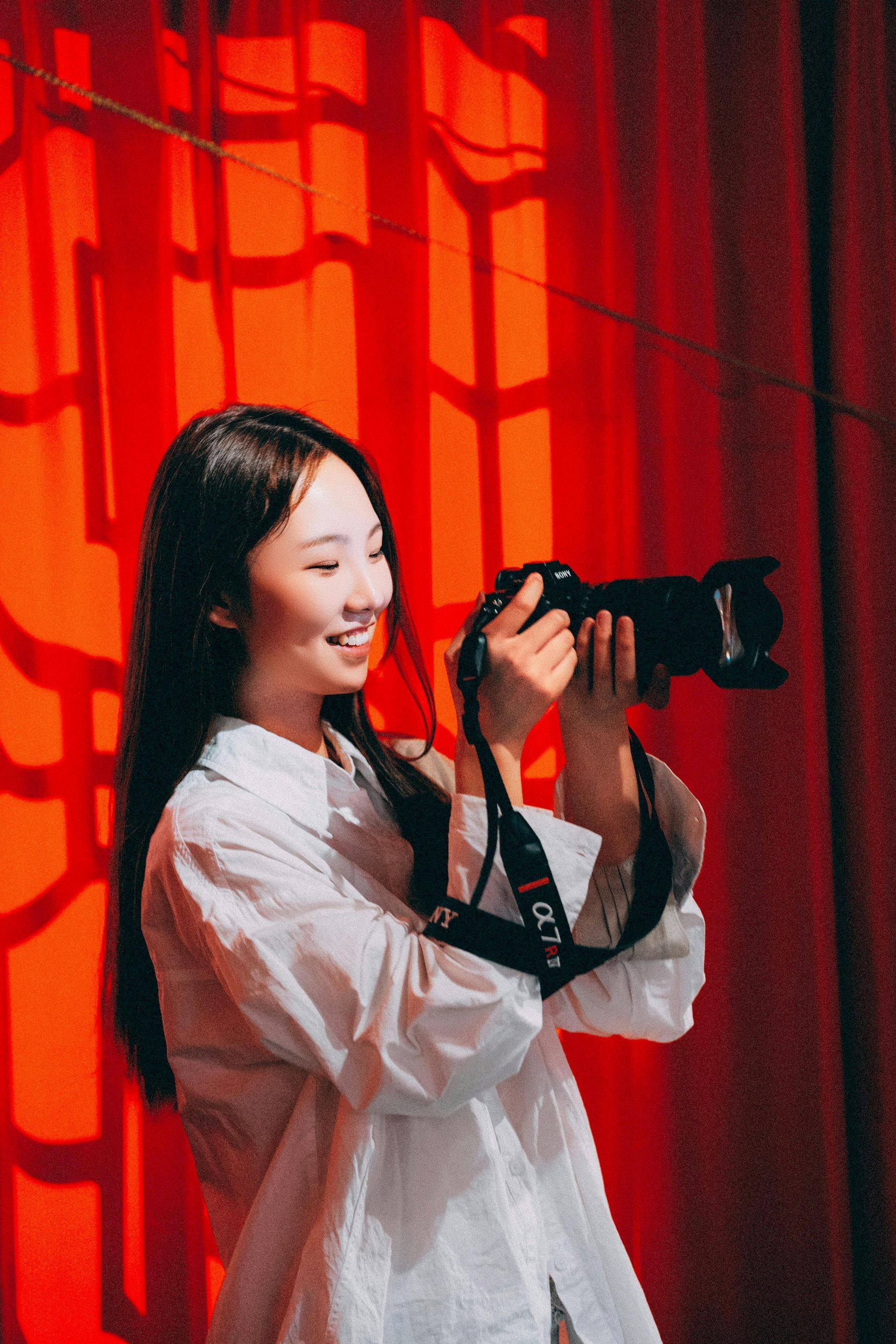 Woman holding a camera, smiling, in front of a red curtain ready to capture a new experience.