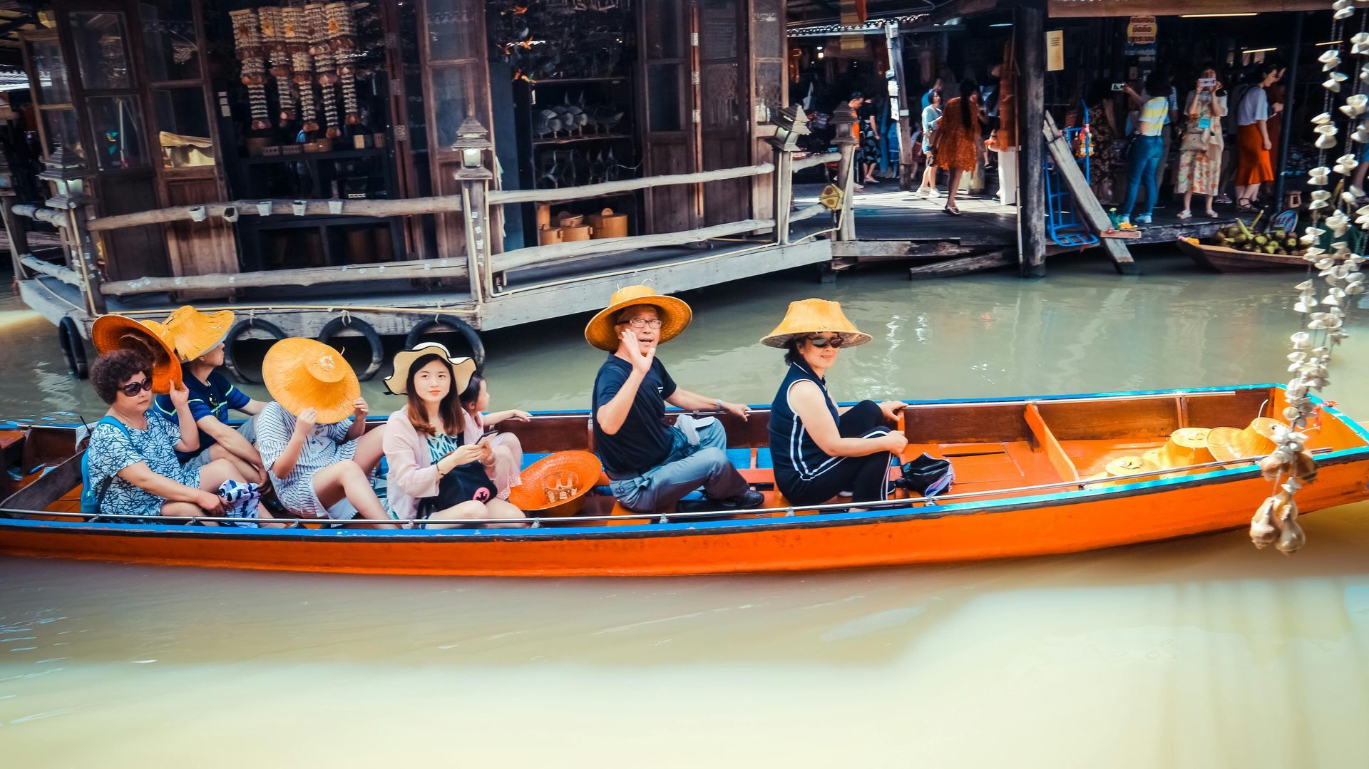 People in straw hats ride an orange boat on a waterway lined with shops and buildings in Asia.